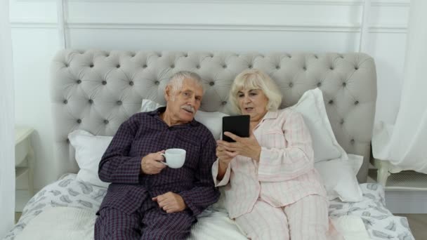 Senior äldre par bär pyjamas liggande på sängen tittar på digitala surfplatta gör online shopping — Stockvideo