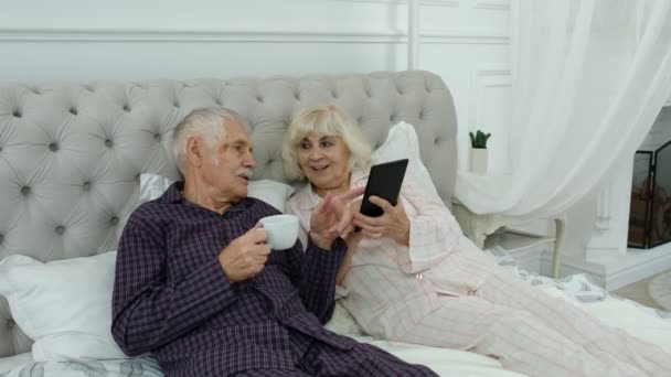 Starší starší pár v pyžamu ležící na posteli při pohledu na digitální tablet dělat on-line nakupování — Stock video