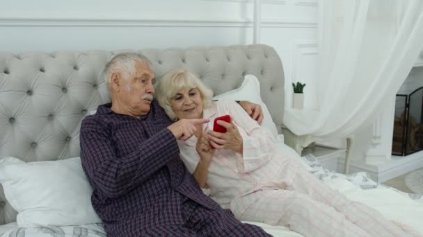 Sénior casal de idosos vestindo pijama deitado na cama olhando para o telefone móvel rindo e se divertindo — Vídeo de Stock