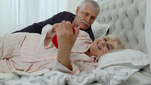 Senior elderly couple wearing pyjamas lying on bed looking on mobile phone laughing and having fun — Stock Video