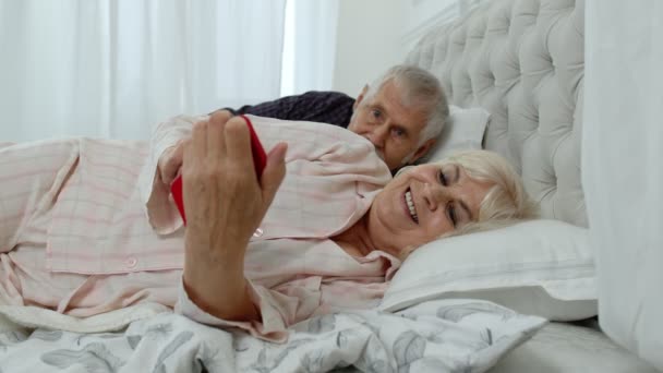 Старшие бабушка и дедушка лежали в постели. Женщина нервничает, что мужчина шпионит за мобильным телефоном — стоковое видео