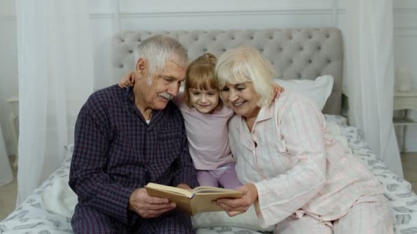 Ragazza carina con anziana nonna in pensione e nonno seduto sul letto lettura libro in camera da letto — Video Stock
