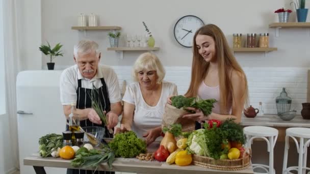 Старшая пара на кухне получает овощи от внучки. Здоровое питание сырой пищей — стоковое видео