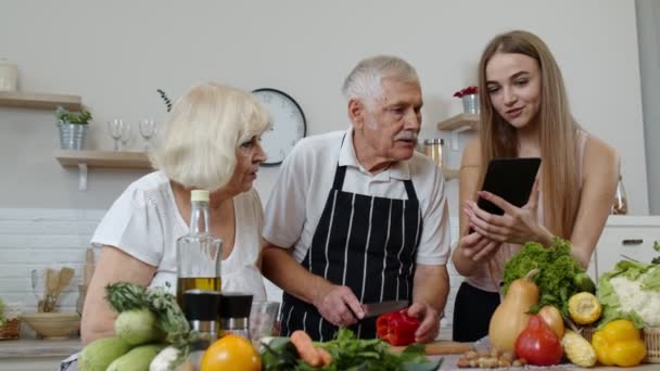 Avós maduros casal cortando legumes para salada, ouvindo receita de menina com tablet — Vídeo de Stock