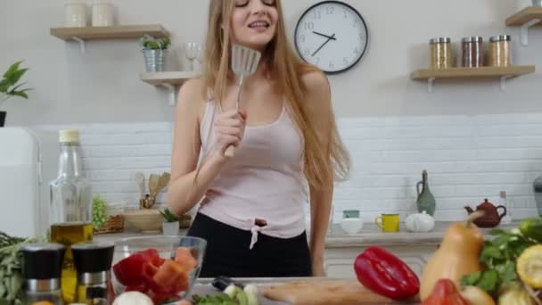 Веселая девушка с длинными волосами танцует и поет на современной кухне. Рацион сырой овощной пищи — стоковое видео