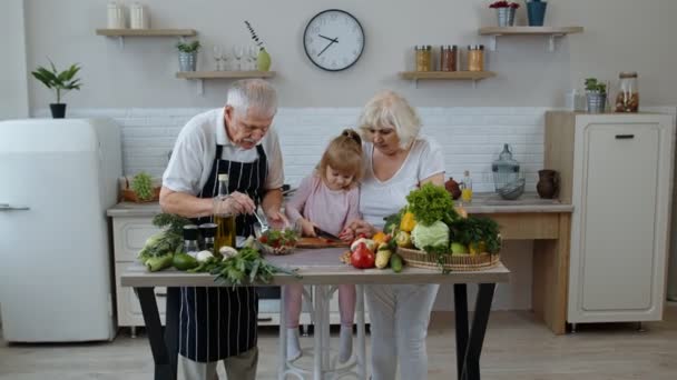 Abuelos ancianos en la cocina enseñando a la nieta a cocinar ensalada, picando pimiento rojo — Vídeos de Stock