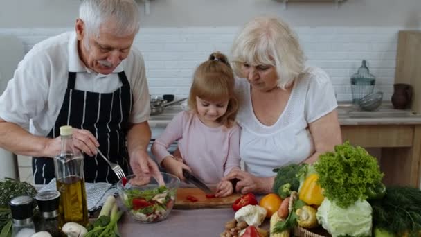 Старша пара на кухні викладає онуку дитині, як готувати, рубаючи перець ножем — стокове відео