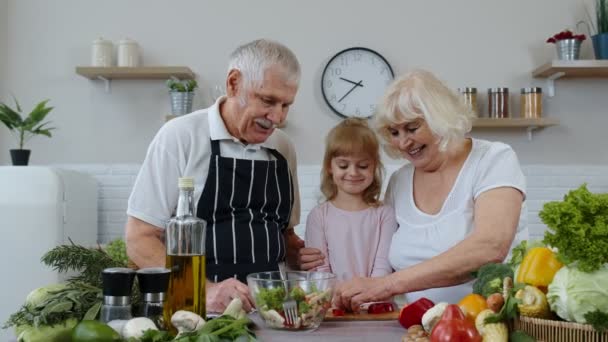 Senior pareja abuela y abuelo en cocina alimentación nieta niño con picado pimienta — Vídeos de Stock