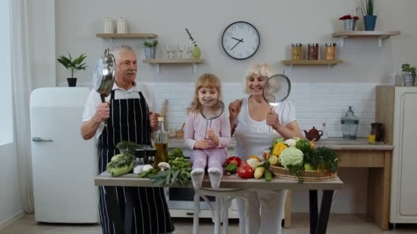キッチンで野菜を調理しながら、幸せなビーガンシニアカップルは孫娘と踊る — ストック動画