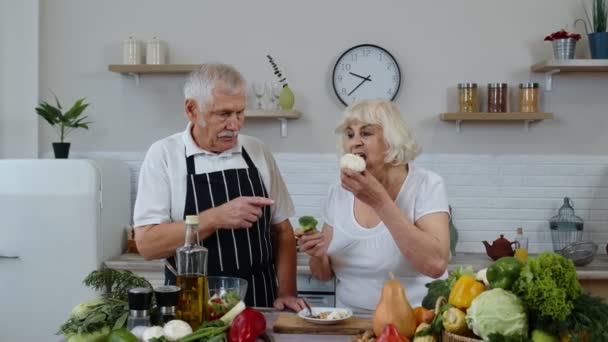 Senior couple in kitchen. Elderly grandmother and grandfather eating raw broccoli and cauliflower — Stock Video