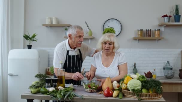Elderly grandparents in kitchen. Funny grandpa joking on grandma. Putting a lettuce about her head — Stock Video