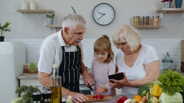 Abuelos mayores pareja con tableta digital y nieta cortar verduras en la cocina — Vídeos de Stock