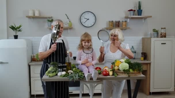 Mulher sênior e homem com neto menina fazendo uma dança engraçada com coador e legumes em casa — Vídeo de Stock