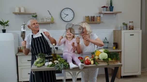 Счастливая веганская старшая пара танцует с внучкой ребенка во время приготовления овощей на кухне — стоковое видео