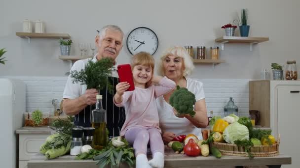 Blogger girl tomando fotos en el teléfono móvil con abuelos mayores en la cocina con verduras — Vídeos de Stock