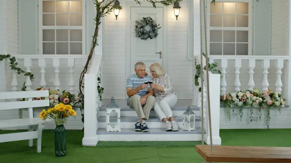 Satisfecho senior ancianos caucásico pareja sentado y beber vino en porche escaleras en casa —  Fotos de Stock