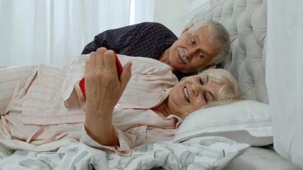 Пожилая пара в пижаме лежит на кровати и смотрит на мобильный телефон, смеясь и веселясь — стоковое фото