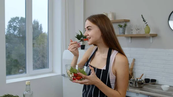Sorridente giovane ragazza mangiare insalata di verdure fresche crude in posa in cucina avendo emozione positiva — Foto Stock