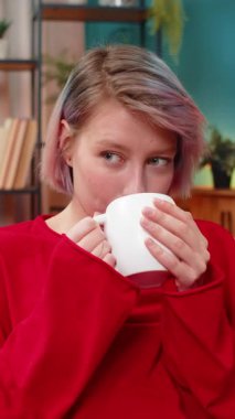 Evde sakince kahve içen genç bir kadın, rahatlamış, tatmin olmuş, huzurlu bir zevk ve rahatlık ifade ediyor. Mutlu kız sessizce gülümsüyor, rahatlıyor, kanepede içecekle keyifli vakit geçiriyor..