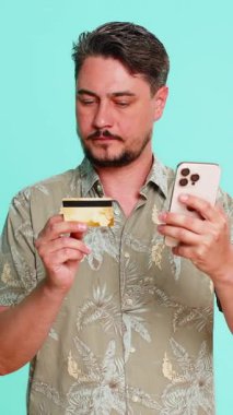 Orta yaşlı adam akıllı telefon ve kredi kartı tutuyor, ödeme düğmesine basıyor çevrimiçi alım-satım yapıyor ya da odaklı ve tatmin edici dijital işlem yapıyor. Mavi arka plandaki genç adam çevrimiçi güvenle alışveriş yapıyor