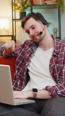 Ekose gömlekli mutlu genç iş adamı yardım hattı web kameralı kulaklıkla konuşuyor ve uzaktan çevrimiçi bir video konferans görüşmesi yapıyor. Kafkasyalı serbest çalışan adam evdeki oturma odasındaki kanepede dizüstü bilgisayar kullanıyor..