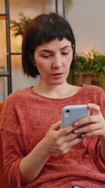 Evdeki kanepede oturmuş akıllı telefon kazanan piyangoyu karıştıran genç bir kadın. Heyecanlı Kafkasyalı kız dairede zafer kutlaması yapıyor yumruklarını sıkıyor. Mutlu bayan çevrimiçi oyun oynuyor