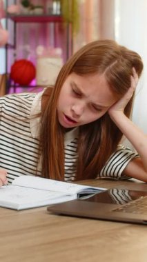 Genç kız, evde kanepede oturuyor, yorgun ve tembel hissediyor, dizüstü bilgisayar ve defterle ödev yapıyor. Okul çocuğu alıştırma kitabına yavaşça yazıyor çevrimiçi öğrenme sırasında odaklanmak için çabalıyor.