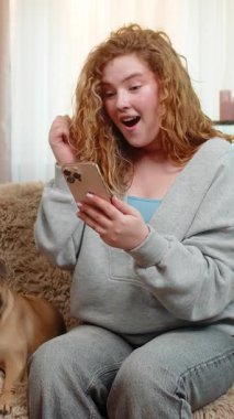 Beyaz saçlı genç bir kadın akıllı telefondan harika haberler okurken kanepesinde sevinç ve heyecanla gülümsüyor. Yanında pug dog olan bir kız var. İkisi de pozitif enerjiyle tepki veriyor..