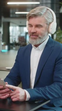 Mutlu, Kafkasyalı olgun iş adamı kulaklık takıyor ve akıllı telefondan favori enerjik disko müziğini dinliyor. Erkek girişimci resmi olarak dinleniyor, ofis lobisinde oturuyor..
