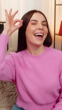 Kanepede oturan mutlu genç bir kadın olumlu bir şekilde gülümseyerek iyi bir jest sergiliyor. Beyaz kız onaylıyor, tatmin oluyor, mutlu oluyor, duygusal olarak anlaşıyor.