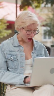 Dizüstü bilgisayarda çalışan neşeli genç bir kadın sevinçle çığlık atıyor zafer jesti olarak ellerini kaldırıyor başarılı oyun paralarını dışarıda kazanıyorlar. Şehirde sandalyede oturan kısa saçlı mutlu sarışın kız.