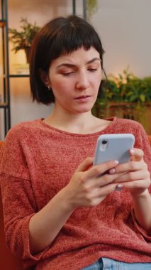 Gündelik giysiler içinde, akıllı telefondan mesaj atan mutlu genç bir kadın. İspanyol kız kanepede oturuyor ve evdeki oturma odasında cep telefonu kullanıyor. Dairede internetten film izleyen bir kadın.