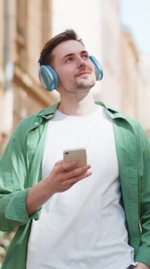 Kablosuz kulaklıklı yakışıklı genç turist dışarıda akıllı telefondan en sevilen enerjik disko müziğini dinliyor. Mutlu Kafkasyalı erkek gezgin, güneşli şehir sokağının önünden geçiyor..