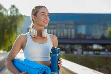 Elinde hasırla genç ve mutlu sporcu kadın parktaki açık hava eğitimine gidiyor. Aktif giyimli, kulaklık takan ve su şişesi kullanan iyimser gülümseyen kız jimnastik eğitimine gidiyor.