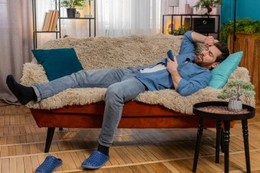 Evdeki genç adam akıllı telefonu elinde tutarken kanepede uyuyakaldı, yorgun hissediyordu. Kafkasyalı adam telefonla dinleniyor, uzun süre ekranda kaldıktan sonra uyanık kalmaya çalışıyor.