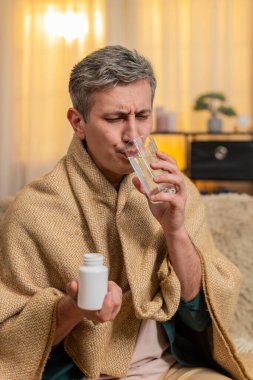 Evdeki genç adam başını tutarken, acı çekerken ve hastalık belirtileri gösterirken hap yutuyor. Kanepedeki beyaz hasta adam yorgun görünüyor ve baş ağrısı, migren ya da fiziksel rahatsızlıkla baş edemiyor.