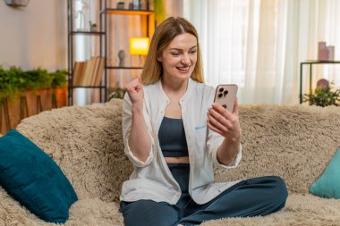 Orta yaşlı bir kadın internet üzerinden ikramiye kazandığını öğrenince sevinçten gülümseyen akıllı telefonları kontrol ediyor. Genç yetişkin kız hareketli zaferi kutluyor, başarıyla mutlu ve mutlu hissediyor.