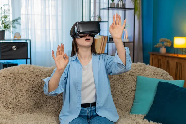 Evde VR kulaklığı takmış, kanepede oturan genç bir kadın inanılmaz bir şekilde başını çeviriyor ve hareketler yapıyor. Beyaz kız heyecan verici bir şekilde sanal gerçekliği keşfediyor, şaşkınlık ve neşe duyguları yaşıyor.