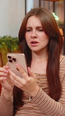 Evdeki genç kadın akıllı telefona bakıyor maç kaybetmenin iç çekişini okuyor üzgün hissediyor ve bakışlarını alçaltıyor. Başarısız bahis sonrası kaşlarını çatan beyaz bir kız stresi hisseder telefonu tutar ve üzgün bir şekilde tek başına oturur.