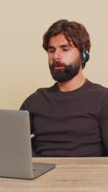 Kulaklık takan genç Hintli işadamı, çağrı merkezi ya da destek operatörü yardım hattı, müşteri ya da iş arkadaşı iletişim desteği ile konuşuyor. Mutlu adam bej renkli stüdyo arka planında masada oturuyor..