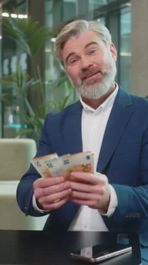 Olgun iş adamı nakit para sayıyor. Avro banknotları kameraya bakıyor, kutluyor, kariyer yapıyor, gelir elde ediyor ve modern ofis lobisinde servetini gösteriyor. Takım elbiseli, kendine güvenen sakallı bir çalışan..