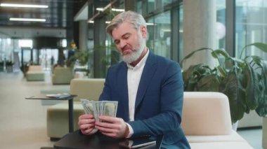 Olgun iş adamı nakit para banknotlarını sayıyor kameralara bakıyor, kutluyor, kariyer yapıyor, gelir elde ediyor, modern ofis lobisinde servet kazanıyor. Takım elbiseli, kendine güvenen sakallı bir çalışan..