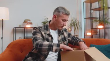 Evdeki kanepede oturan mutlu genç adam teslimat paketini açıyor. Gülümseyen memnun erkek alışverişci çevrimiçi dükkan müşterisi karton kutu açıyor hızlı posta yoluyla satın alma hediyesi alıyor.