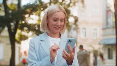 Portre olgun bir iş kadını internette cep telefonuyla dışarıda sohbet ediyor. Mutlu gülümseyen orta yaşlı başarılı kadın cep telefonu kullanıyor. Şehir caddesinde SMS ile iletişim kuruyor.