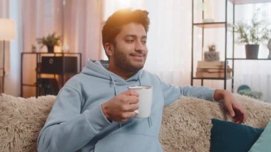 Gülümseyen Hintli adam evde sıcak kahve ya da bitki çayı içiyor. Hindu genç adam moladan zevk alıyor, sabahları kanepede oturuyor, düşünceli bir şekilde uzaktan bakıyor ve dinleniyor.