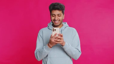 Mutlu Hintli genç adam sosyal medyada geziniyor, akıllı telefona bakarken gülümsüyor, ilginç içeriğin tadını çıkarıyor ve bir mesaj yazıyor. Çevrimiçi bağlanan rahat Arap adam, pembe arka planda izole edilmiş.