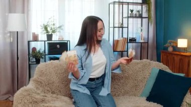 Evdeki genç mutlu kadın kanepenin üzerinde sevinçle dans ediyor elinde nakit avroluk banknotlar tutarak piyango kazanmasını kutluyor. Beyaz kız heyecanını, finansal başarısını, zenginliğini ifade etmekten mutlu.