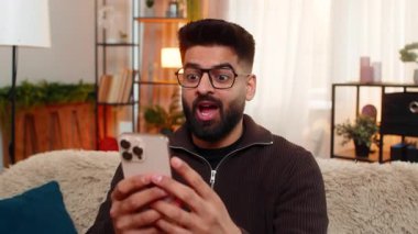 Evdeki Hintli adam gözlerini kocaman açıyor ve akıllı telefona bakarken kaşlarını kaldırıyor. Şok edici bir mutluluk tepkisiyle. Kanepedeki Arap adam el hareketleriyle, beklenmedik iyi bir mesaj karşısında şaşırdı..