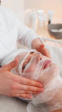 Orta yaşlı bir kadın kozmetoloji kliniğinde yüz bakımı yapıyor. Güzellik uzmanı karboksiterapi sırasında dikkatli bir şekilde gazlı bez maskesi takar. Yenilenme, ışıltı, esneklik ve derin su kaybına odaklanır.