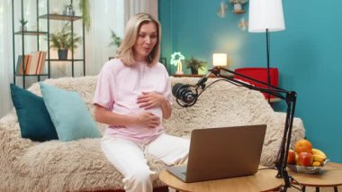 Hamile, orta yaşlı bir kadın dizüstü bilgisayarı ve stüdyo mikrofonuyla oturup anneler için bahşiş veriyor. Kanepede oturan beyaz kız görüntülü konuşma sırasında gülüyor..